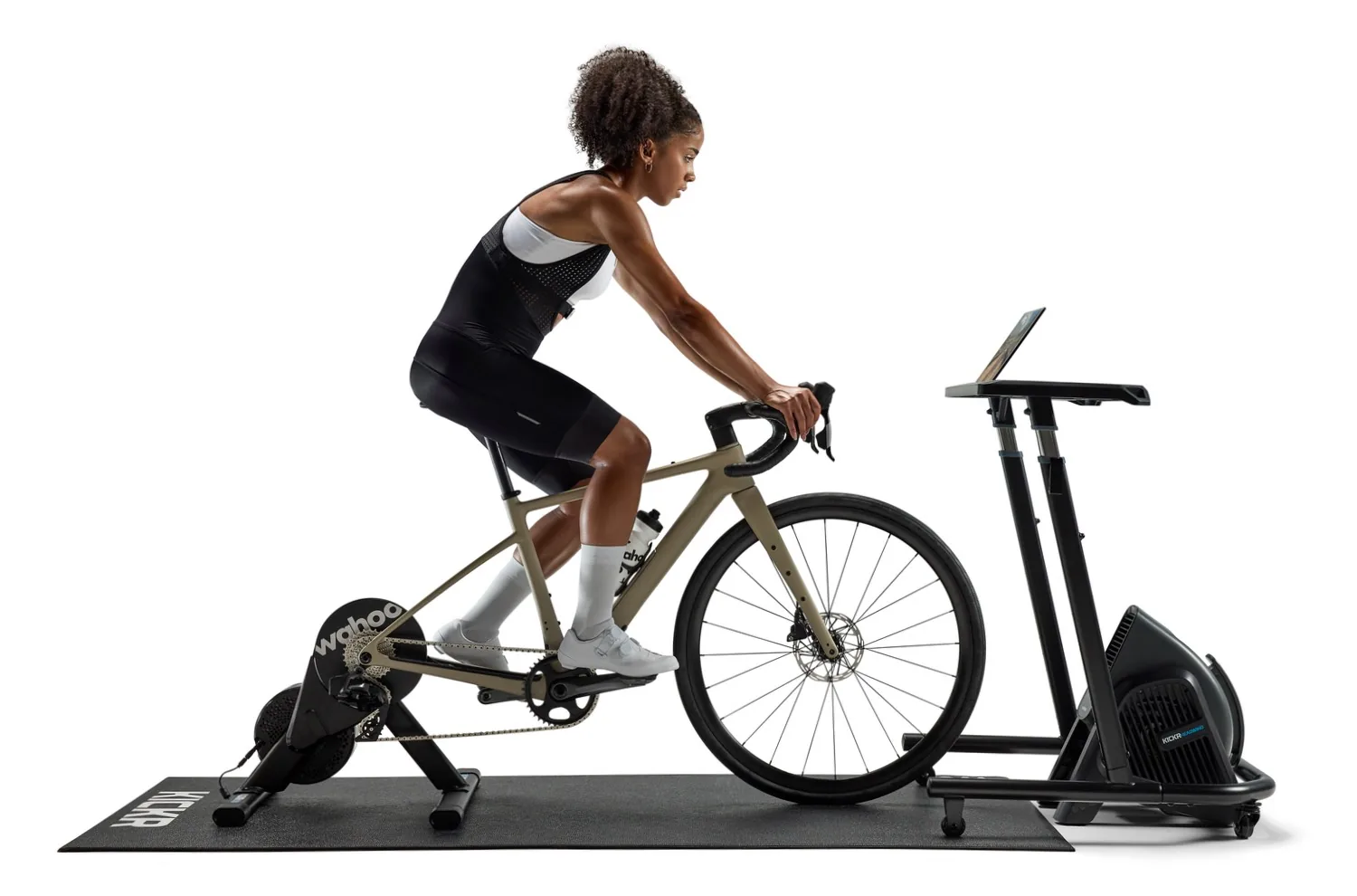 Indoor cycling setup with Wahoo accessories including heart rate monitor, KICKR desk, HEADWIND fan, CLIMB grade simulator, pedals, and mat